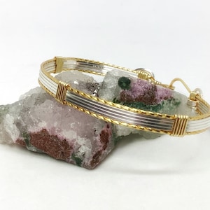 May include: A silver and gold wire wrapped bangle bracelet. The bracelet is made of two different metals, silver and gold, and is wrapped in a wire design. The bracelet is on a white background.