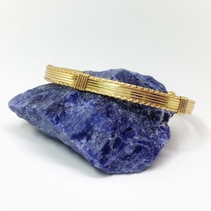 Puede incluir: Una pulsera de brazalete de alambre en tono dorado con múltiples hilos paralelos y dos bandas decorativas envueltas. La pulsera descansa sobre una piedra de sodalita azul profundo rugosa con texturas cristalinas e inclusiones blancas.