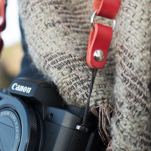 May include: A black Canon camera with a zoom lens and a red camera strap with a silver buckle. The camera is partially covered by a gray and white knitted scarf.