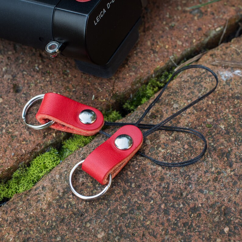May include: Two red leather camera straps with silver metal rings and a black cord. The straps have a silver metal button.