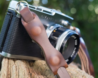 May include: A silver camera with a brown leather camera strap. The strap has a decorative stitching detail and a metal ring.