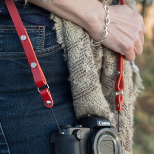 May include: A red leather camera strap with silver metal accents and a Canon camera with a black lens.