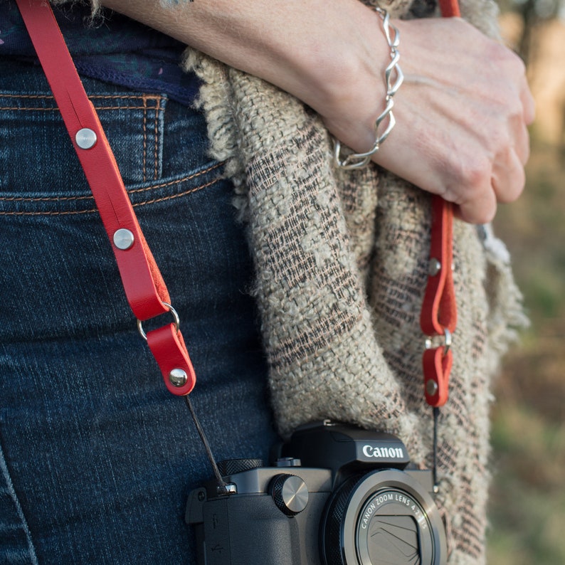 May include: A red leather camera strap with silver metal accents and a Canon camera with a black lens.