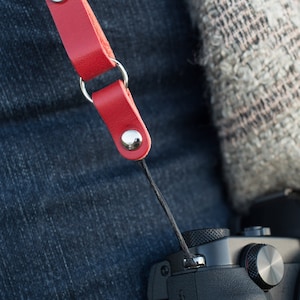 May include: A red leather camera strap with silver metal hardware and a black cord.