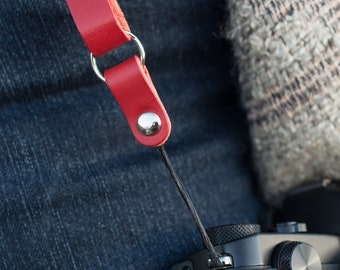 May include: A red leather camera strap with silver metal hardware and a black cord.