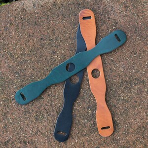 May include: Three leather camera straps in different colors: dark green, black, and brown. The straps have a hole in the middle and are designed to be adjustable.