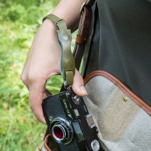 May include: A person wearing a green shirt and a brown leather camera bag with a green camera strap. The camera bag has a zipper and a flap. The camera strap has a metal buckle. The person is holding a black Olympus Pen F camera with a red lens. The camera has a silver lens ring and a black strap.