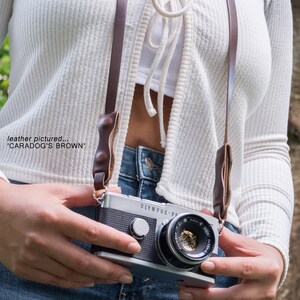 May include: A silver Olympus Pen F camera with a brown leather camera strap. The camera has a black lens with a silver ring around it. The strap has a brown leather pad on each side.