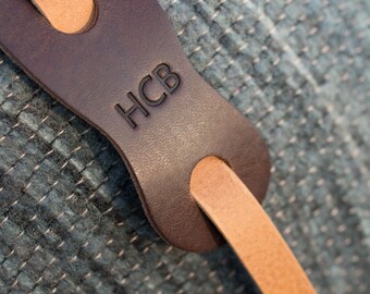 May include: Close-up of a brown leather strap with a tan leather loop. The strap has the letters "HCB" embossed on it.