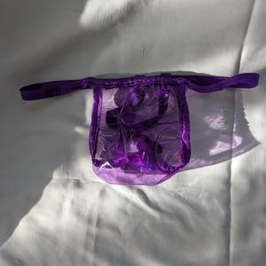 Könnte beinhalten: Ein transparenter, violetter String-Tanga mit passendem elastischen Bund. Der String besteht aus einem klaren, flexiblen Material und ist minimalistisch gestaltet. Das Kleidungsstück liegt auf einer weißen Oberfläche.
