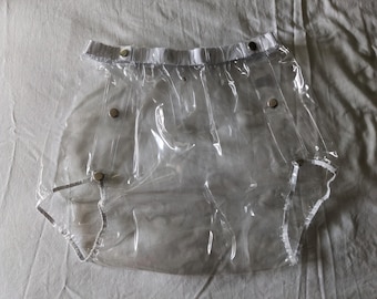 May include: Clear plastic waterproof garment with snap closures. The garment has a white waistband and leg openings. The material is transparent, allowing the underlying clothing to be visible. The garment is designed for protection.