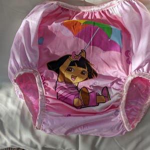 May include: Pink waterproof training pants featuring a cartoon character holding a colorful umbrella. The pants have elastic waist and leg openings. The design includes a cartoon character in a pink outfit.
