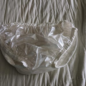 May include: A white, plastic diaper cover with a ruffled edge and white polka dots. The cover is made of a shiny, waterproof material and is designed to be worn over a diaper. The diaper cover is laying on a white, textured surface.