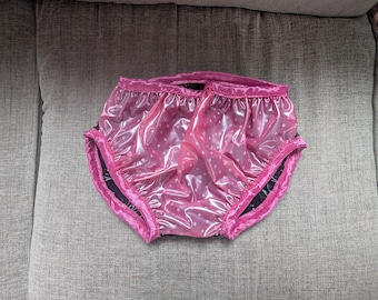 May include: Pink plastic bloomers featuring a white polka dot pattern. The garment has a shiny, reflective surface with gathered elasticized openings at the waist and leg holes, finished with a matching pink fabric trim along the edges.