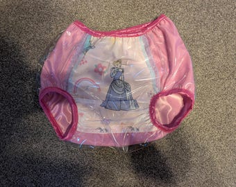 May include: Pink plastic nappy cover featuring a central graphic of a princess in a blue gown, surrounded by small floral and rainbow motifs. The garment has bright pink elasticated leg openings and a matching pink waistband, encased in clear plastic.