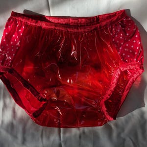 Pode incluir: Cuecas de plástico vermelhas com bolinhas brancas nas laterais. A roupa interior tem cós elástico e aberturas para as pernas. O material parece brilhante e translúcido, com dobras e vincos visíveis.
