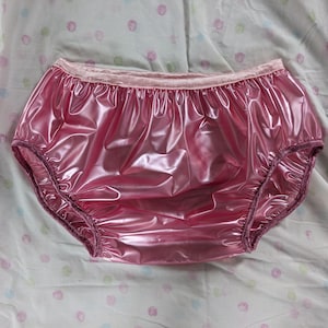 Puede incluir: Bragas de plástico rosa metálico brillante con cintura elástica fruncida y aberturas para las piernas. La prenda tiene una superficie lisa y reflectante con un acabado de alto brillo, un diseño sencillo y bordes con volantes en la cintura.