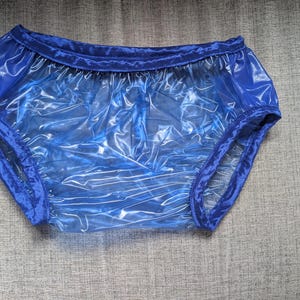 May include: A pair of translucent blue plastic bloomers featuring a shiny, crinkled texture. The garment has a gathered elastic waistband and leg openings, both finished with a matching solid blue fabric trim along the edges.