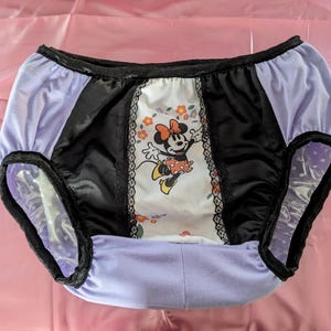 Puede incluir: Bragas lavanda y negras con un diseño de Minnie Mouse. El panel central muestra a Minnie Mouse con detalles florales. Los bordes están adornados con encaje negro y tela de satén negro.