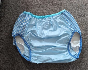 May include: High-waisted plastic pants featuring a solid light blue centre panel and side panels with a blue and white gingham check pattern. The garment has a bright turquoise elastic waistband and blue elastic trim around the leg openings.