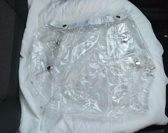 May include: Clear plastic waterproof pants featuring a high-waisted design with a white waistband and silver-toned metal snap buttons along the front and leg openings. The garment is constructed from a transparent, flexible material with reinforced seams.