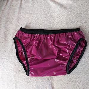 Pode incluir: Uma cueca de cor magenta com um acabamento brilhante e reflexivo. A peça apresenta um cós elástico preto sólido e acabamento preto ao longo das aberturas das pernas. O material parece liso e brilhante, com tecido franzido nas costuras.
