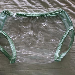 May include: Clear plastic underwear with mint green trim. The underwear has a classic brief style with elasticized leg openings and waistband. The material appears to be waterproof and the trim has a satin sheen.