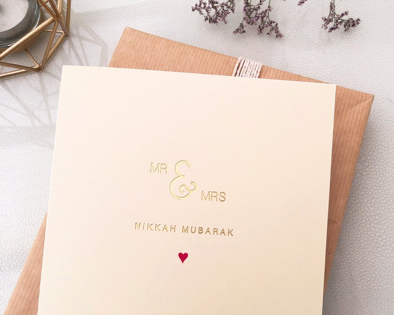 May include: A cream-colored card with gold lettering that reads "Mr & Mrs Nikkah Mubarak" with a small red heart at the bottom.