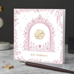 Può includere: Biglietto di auguri quadrato con sfondo bianco e bordo floreale rosa. Il biglietto presenta un arco rosa con calligrafia araba dorata e le parole "EID MUBARAK" in oro. Stelle dorate sparse sulla carta.