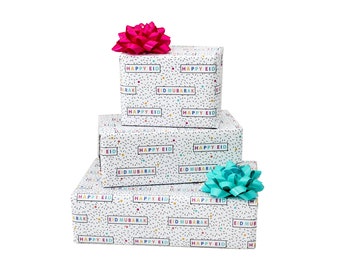 May include: Three gift boxes stacked on top of each other, wrapped in white paper with a colorful confetti pattern and the words "Happy Eid" and "Eid Mubarak" printed on them. The top box has a pink bow, the middle box has a turquoise bow, and the bottom box has a turquoise bow.