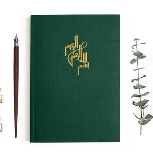 Pode incluir: Um caderno verde escuro com uma inscrição dourada em escrita árabe. O caderno está sobre uma superfície branca com uma caneta de caligrafia marrom e dois clipes de papel dourados.