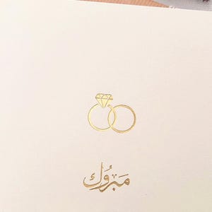 Peut inclure: Une carte blanche avec des lettres dor&eacute;es et une illustration dor&eacute;e de deux anneaux entrelac&eacute;s avec un diamant au sommet. Le texte sur la carte est "Mabrouk".