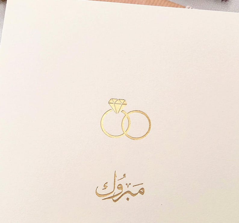 Peut inclure: Une carte blanche avec des lettres dor&eacute;es et une illustration dor&eacute;e de deux anneaux entrelac&eacute;s avec un diamant au sommet. Le texte sur la carte est "Mabrouk".