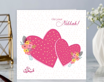 May include: A pink and white card with two pink hearts with gold glitter and a floral design. The card says "On your Nikkah!" and has Arabic text that says "Mabrook".