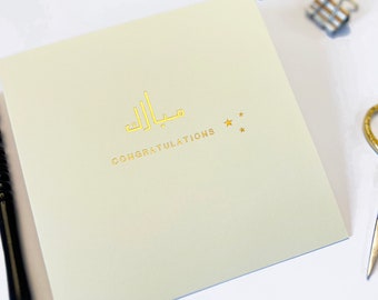 Può includere: Un biglietto verde chiaro con lettere dorate in rilievo che dicono "Congratulazioni" e "Mabrook" in scrittura araba. Ci sono tre stelle dorate a destra del testo arabo.