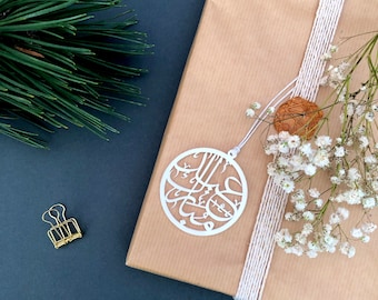 May include: A white, circular ornament with Arabic calligraphy hangs over a wrapped gift. The gift is wrapped in brown paper and tied with white twine. A sprig of white flowers and a gold paper clip are also present.
