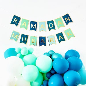 May include: A blue and green banner with gold lettering that reads "Ramadan Mubarak" hangs above a collection of blue and green balloons.