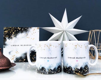 May include: Two white ceramic mugs with black and gold marble designs. The mugs have the text "Umrah Mubarak" with a blue star on one mug and a pink heart on the other mug.