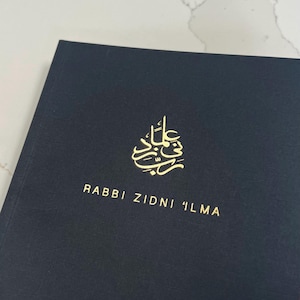 Pode incluir: Um caderno preto com letras douradas que dizem "Rabbi Zidni 'Ilma".