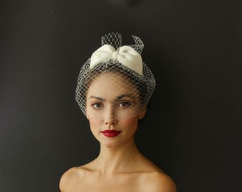May include: A vintage-style headpiece featuring a white bow and a delicate net veil. The headpiece is set against a dark background. The woman in the image is wearing a black off-the-shoulder top and red lipstick.