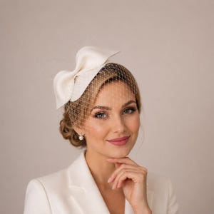 May include: A white fascinator hat featuring a structured bow design and a delicate birdcage veil that covers the upper portion of the face. The headpiece is crafted from smooth fabric and is paired with a matching white blazer, creating a cohesive look.