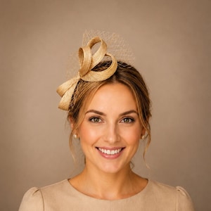 Op de afbeelding: Een beige fascinator met gestructureerde, geluste stoffen linten en een fijn, bijpassend netje. Het accessoire heeft een geweven, textuurrijke uitstraling en is aan één kant geplaatst, wat zorgt voor een gelaagd en sculpturaal effect.