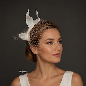 Op de afbeelding: Een witte fascinator-hoofdtooi met een gestructureerde strik met twee rechtopstaande lussen en een delicate birdcage-sluier van fijn gaas. Het accessoire is aan de zijkant van het hoofd geplaatst, met de tekst Feltpoint zichtbaar in de linkerbenedenhoek.