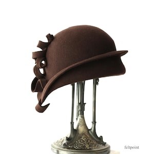 Peut inclure: Un chapeau cloche en feutre marron avec un nœud décoratif sur le côté. Le chapeau a une couronne arrondie et un bord légèrement relevé. Le chapeau est présenté sur un présentoir à chapeau argenté.
