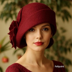 Peut inclure: Un chapeau cloche bordeaux avec une grande fleur sur le côté. Le chapeau en feutre a une couronne arrondie et un bord relevé. L'image montre également le texte "Feltpoint".