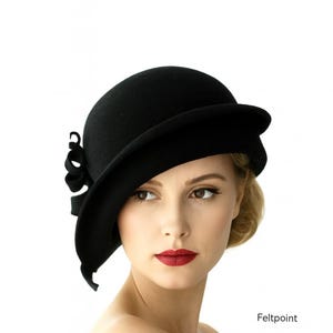 May include: A black cloche hat with a wide brim and a decorative bow on the side. The hat is made of felt and has a classic, vintage design. The hat is a stylish accessory.