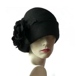 Pode incluir: Um chapéu cloche de feltro preto com um grande adorno de feltro preto em forma de flor. O chapéu está inclinado para baixo sobre a testa.