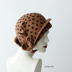 May include: A tan cloche hat with dark brown polka dots. The hat features a decorative, curled fabric accent on the side. The brim is slightly turned up. The hat is displayed on a white mannequin head.