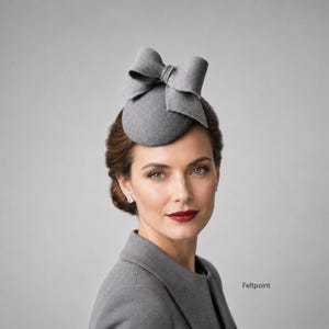 May include: A grey felt fascinator hat featuring a large, structured bow detail on top. The hat is dome-shaped and sits atop the head. The word Feltpoint is printed in the lower right corner of the image, which shows a grey garment as well.