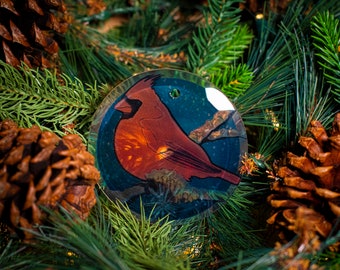 May include: A round, clear glass Christmas ornament featuring a detailed illustration of a red cardinal bird. The bird is perched on a branch against a dark blue and green background. The ornament is surrounded by green pine needles and brown pine cones.